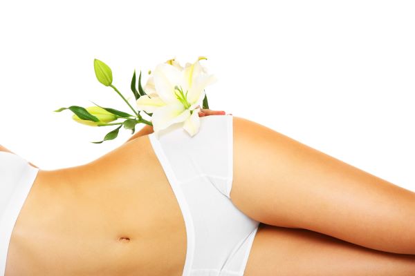 Close da metade do corpo, lindo, de uma mulher deitada de lado. Ela veste lingerie branca e, sobre a calcinha, há um ramo de lírio branco. 
Conceito de rejuvenescimento íntimo por meio do uso do laser.