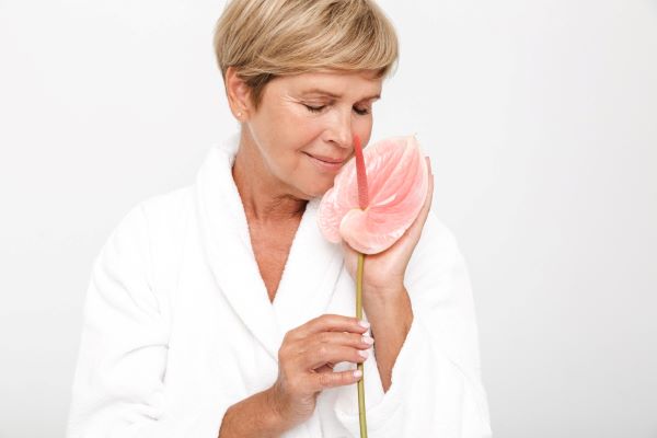 Mulher adulta e sensível com cabelo loiro curto e roupão branco segurando uma flor rosa junto ao rosto.