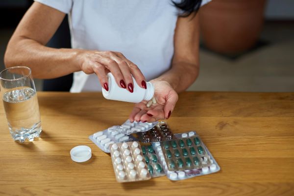 Metade do corpo de uma mulher que segura um pote de comprimidos de reposição hormonal oral, virando-os sobre a mão. Sobre a mesa há um copo de água e várias cartelas de outras medicações.