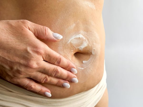 Close-up da mão da mulher com unhas brancas aplicando creme de reposição hormonal transdérmica no abdomen. 