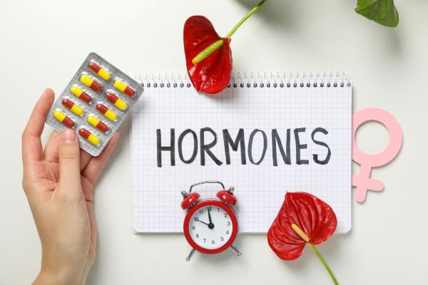 Tratamento com medicamentos hormonais. Imagem com um caderno onde está escrito HORMONES e sobre o qual estão 2 antúrios vermelhos, 1 relógio despertador vermelho e uma mão feminina segurando um blister de medicação.