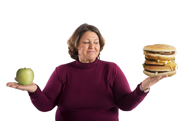 Mulher obesa de meia idade segura uma maça verde numa das mãos e na outra um hambúrguer grande. 
sua expressão demonstra dúvida sobre a alimentação.
