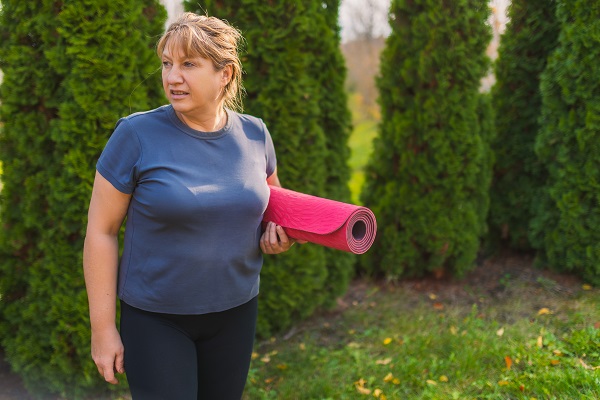Mulher de meia idade, vestindo roupas esportivas e segurando um tapete de yoga, caminha pelo parque.