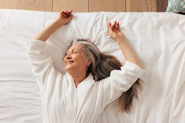 Mulher madura, com cabelos compridos e grisalhos, vestindo roupão branco e deitada sobre uma cama, sorri feliz. 